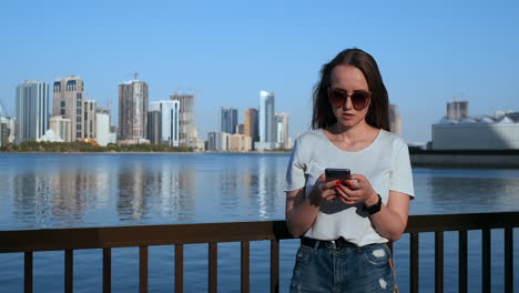 Hermosa-Chica-Con-El-Pelo-Largo-Con-Gafas-De-Sol-Usando-Una-Aplicación-Para-Teléfonos-Inteligentes-En-El-Muelle-Del-Río-Sunset-Cerca.