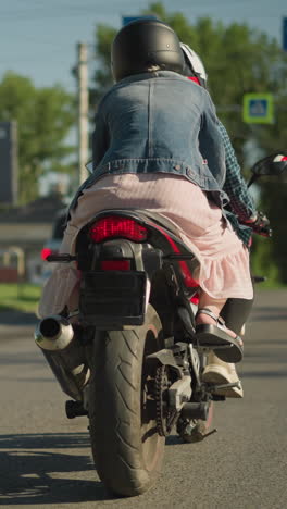 dos mujeres montando una bicicleta eléctrica se acercan lentamente a un semáforo mientras los coches esperan la luz verde, el amigo del jinete se aferra por detrás, se ve un borrón de edificios, señales y coches a su alrededor