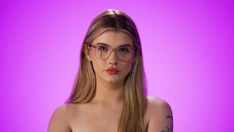 medium shot of a young pretty blonde woman wearing glasses and touching them with her hands while looking into the camera in front of purple background