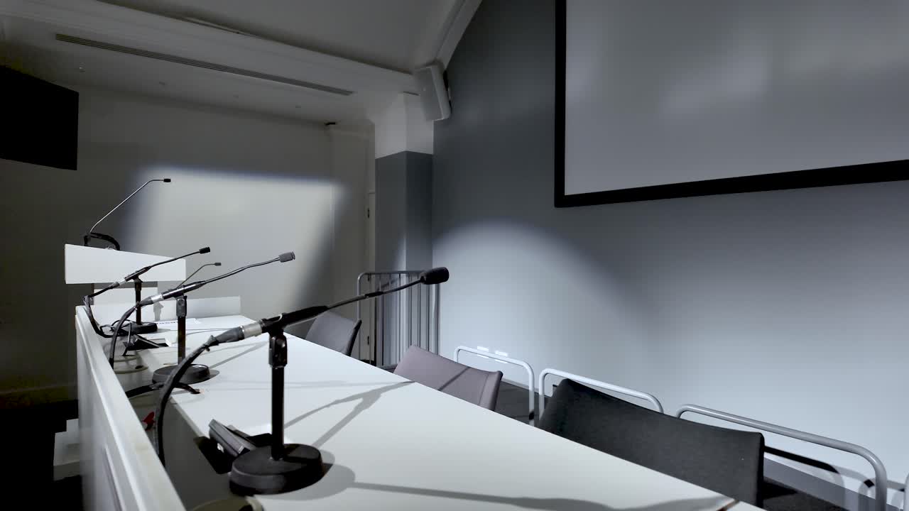Sala conferenze vuota con microfoni sul tavolo bianco, schermo di presentazione e sedie per eventi aziendali, seminari e impegni pubblici