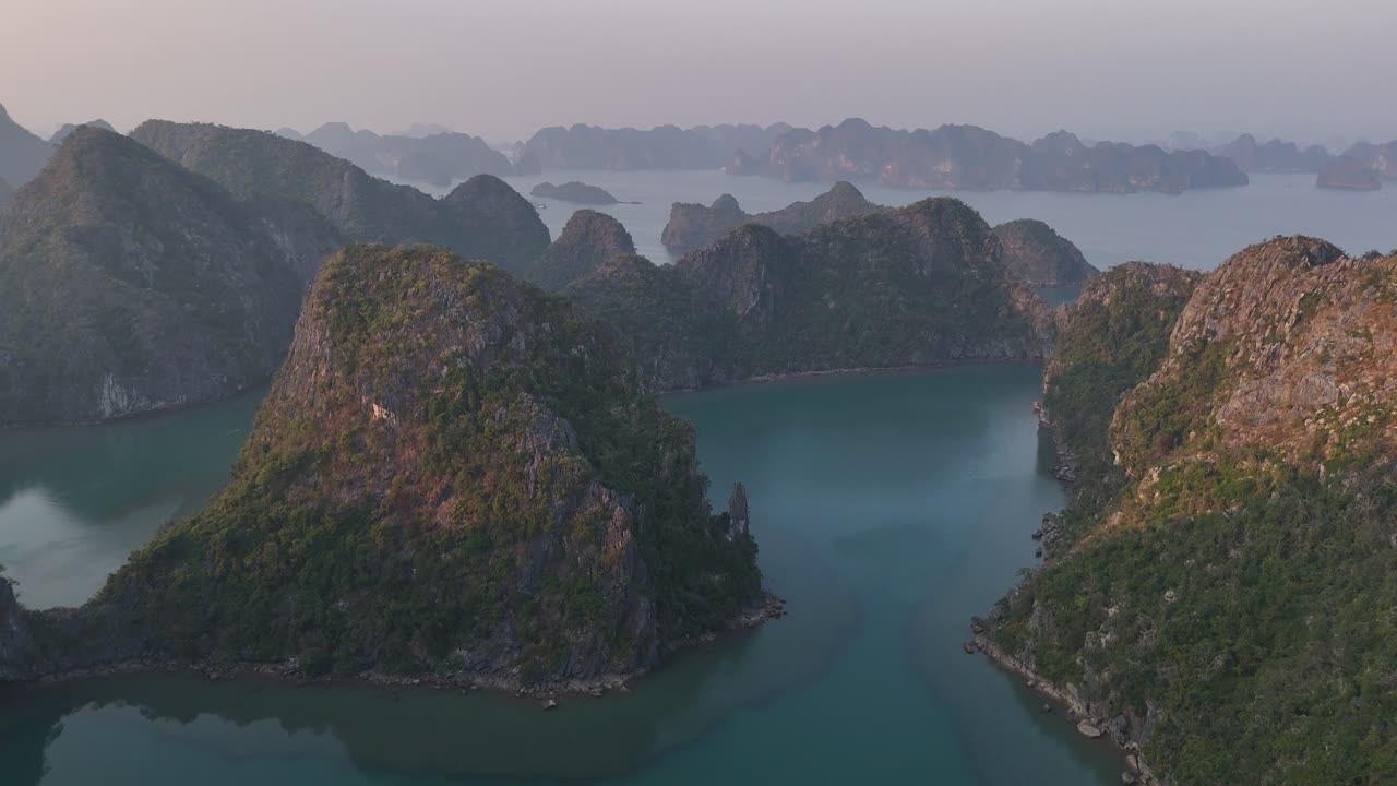 Aerial drone view of peaceful bay surrounded by steep forested mountains at dawn, with soft morning light casting shadows over calm turquoise water in remote and dramatic coastal terrain