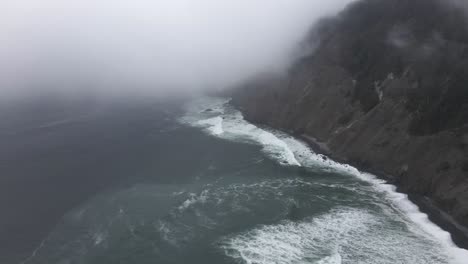 fog hangs tight to steep cliffs as waves crash along a jagged coastline, aerial