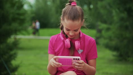 a young girl enjoying her time outdoors while engaged with a pink electronic device, wearing matching pink headphones and a relaxed expression, surrounded by lush greenery