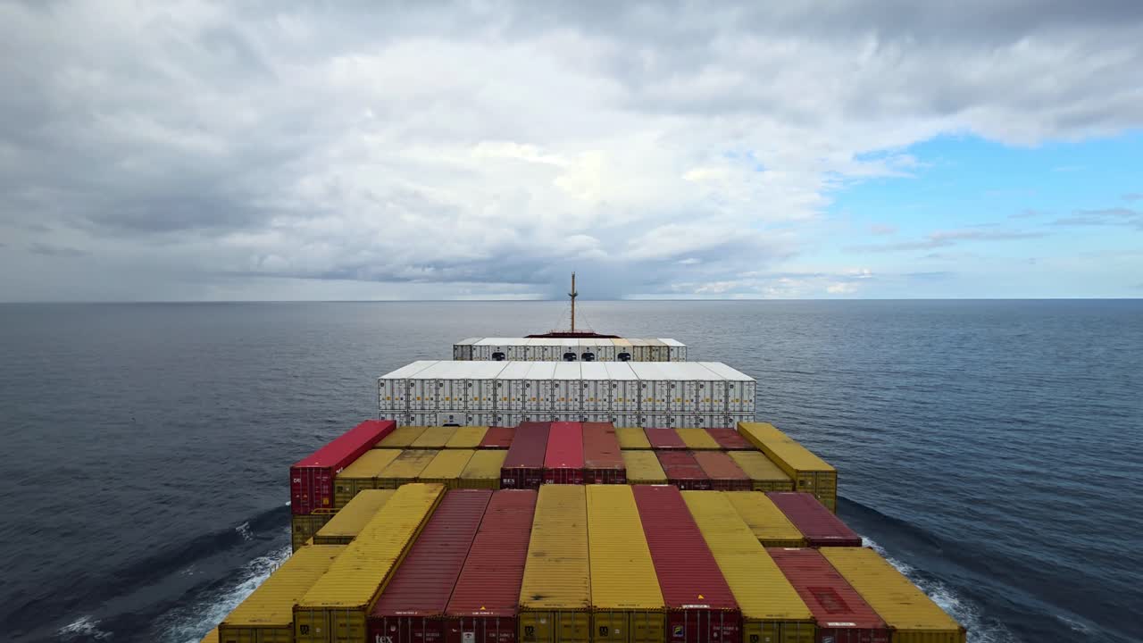 Luftfoto i fugleperspektiv af et fuldt lastet containerskib, der sejler gennem åbent hav under en overskyet himmel.