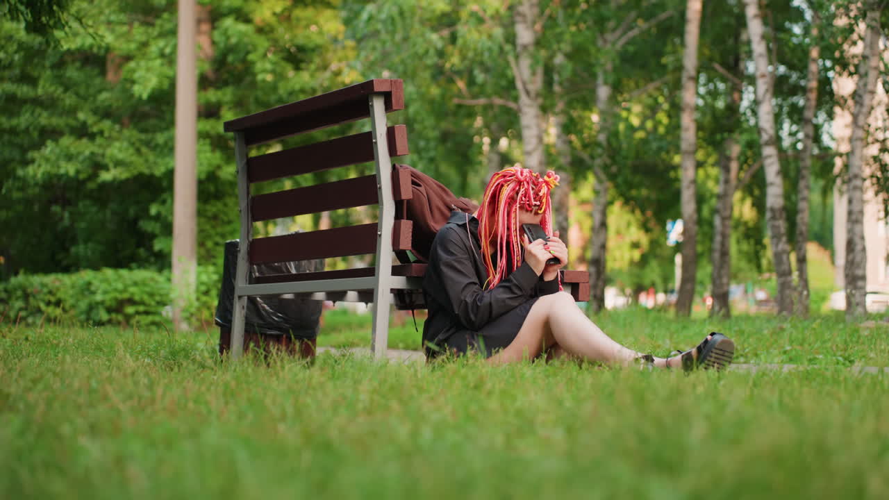 잎이 무성한 공원의 벤치 옆에 앉아 다리를 뻗고 풀밭에 앉아 스마트폰을 보며 먼 곳을 응시하는 핑크색 드레드락을 한 백인 여성, 편안함과 성찰을 추구하는 분위기