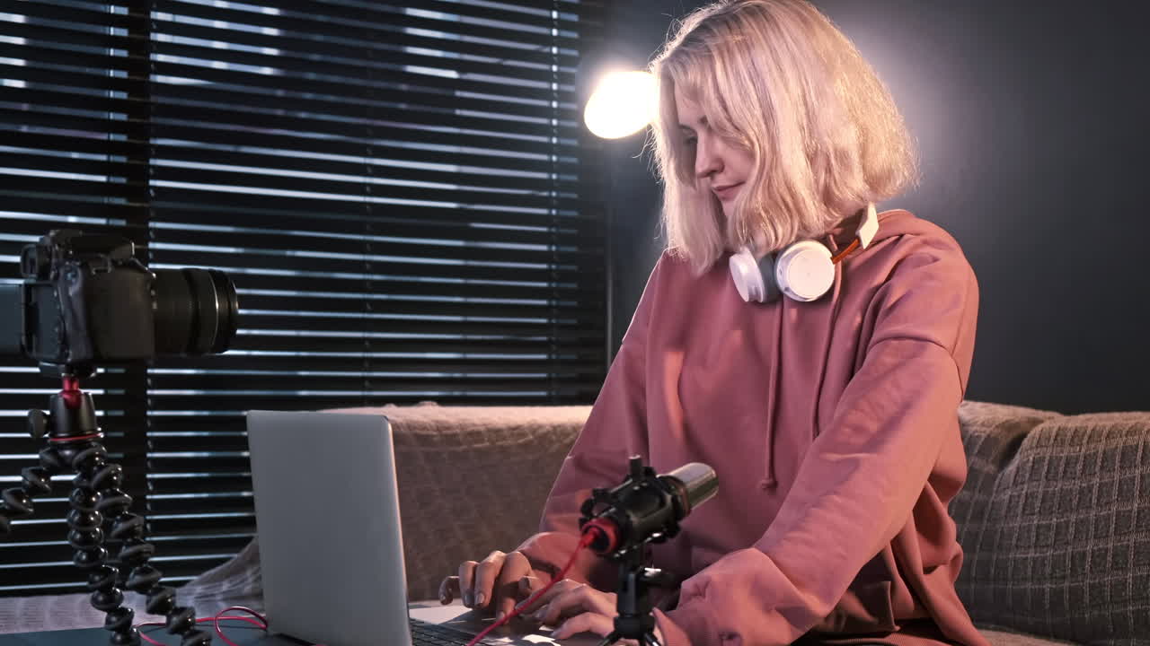 Young content creator blonde girl with headphones working on her laptop on the table with camera on a tripod, microphone. Working from home