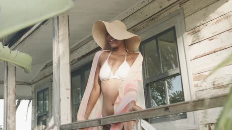 Biracial-woman-in-bikini-and-sunhat-smiling-on-porch-of-wooden-beach-house,-slow-motion