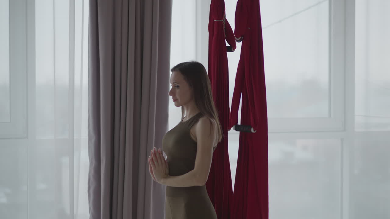 Side view spiritual mentor practicing quietness in spa overlooking red silk hammocks wearing brown leggings focusing on mindfulness balance under soft ambient light reflecting inner calm presence