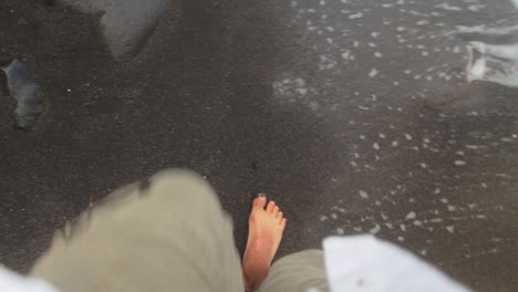 Aufnahme-Aus-Der-Perspektive-Eines-Mannes,-Der-Barfuß-An-Einem-Schwarzen-Sandstrand-Läuft,-Während-Wellen-Auf-Seine-Lauffüße-Schlagen