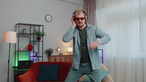 overjoyed young man in wireless headphones dancing, singing on cozy couch in living room at home