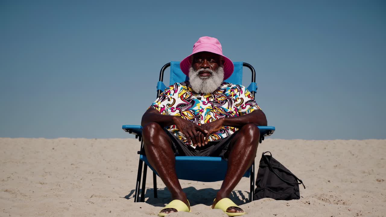 Fashionable senior man with a white beard and a pink bucket hat sits comfortably on a blue beach chair on a sunny beach, enjoying his summer vacation with a relaxed expression