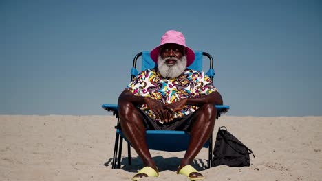 fashionable senior man with a white beard and a pink bucket hat sits comfortably on a blue beach chair on a sunny beach, enjoying his summer vacation with a relaxed expression