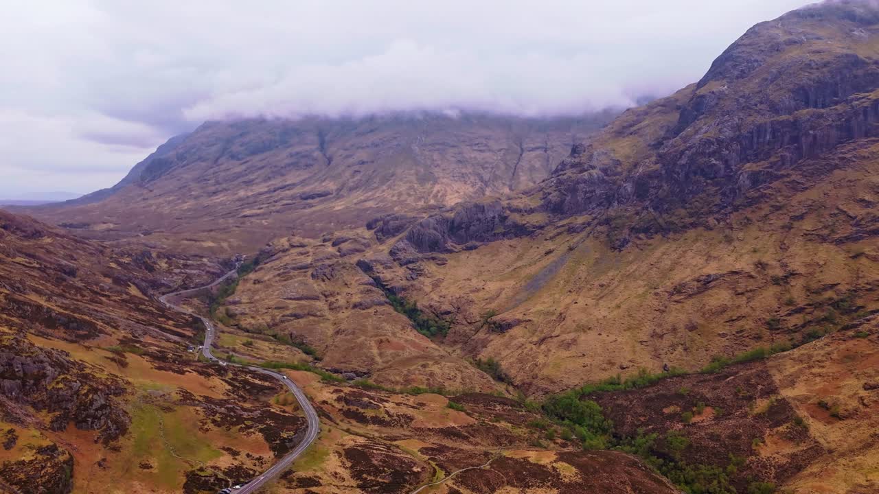 Ripresa aerea con drone, in avvicinamento, su Glencoe in Scozia che rivela montagne spettacolari, una strada tortuosa e un paesaggio di valle nebbioso sotto cieli nuvolosi