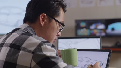 close up side view of asian male drinking coffee while drawing new car design concept on a tablet in the studio with tv and computers display 3d electric car model