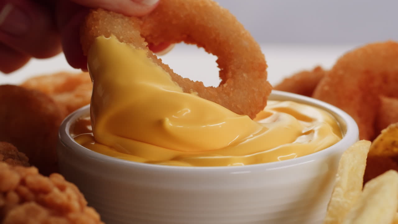 Hand dipping onion ring in cheese sauce with other fast food