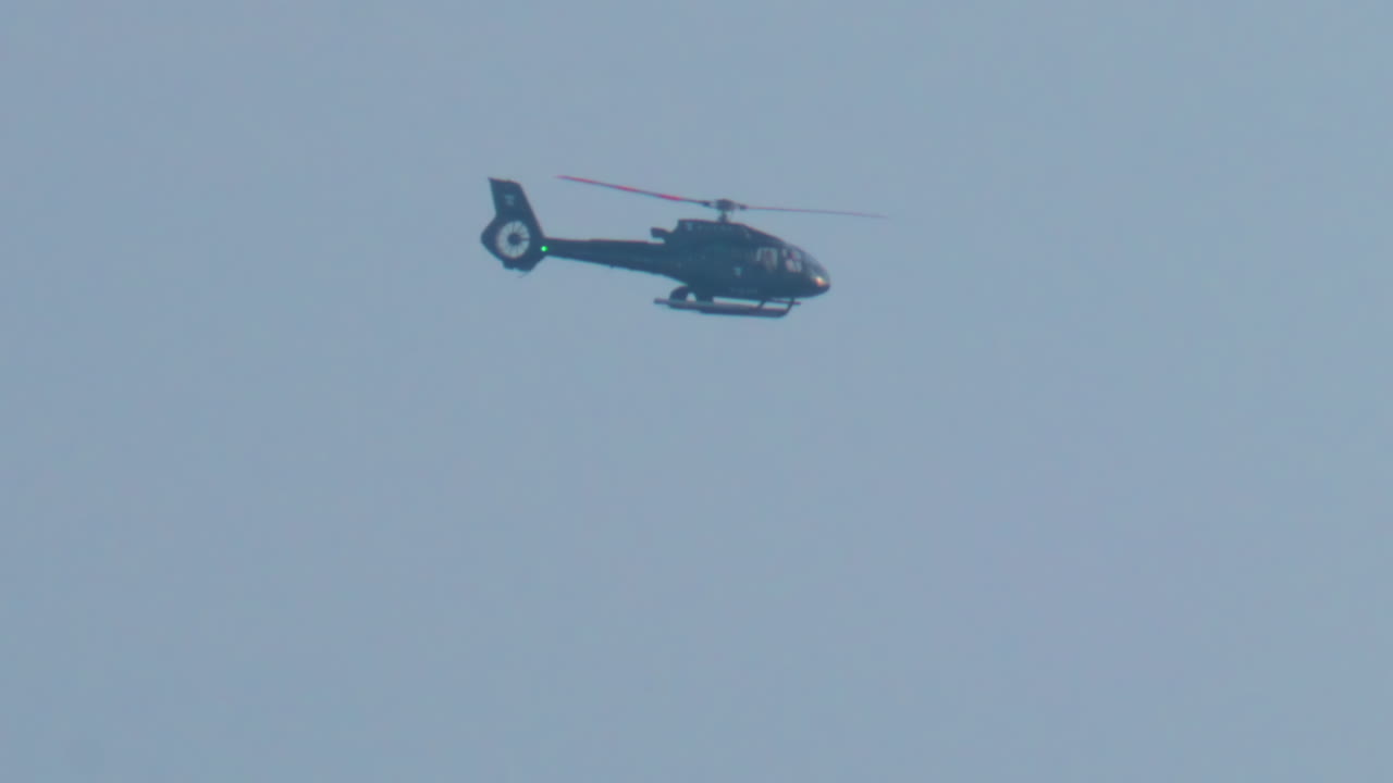 A black helicopter flies across a clear blue sky in bright daylight