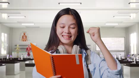 close up of asian teen girl student with a backpack reading book and screaming goal celebrating succeed learning in science laboratory