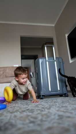 a black cat stands near the grey suitcase. baby boy hiding behind the suitcase appears and crawls to the camera scaring the cat. vertical video.