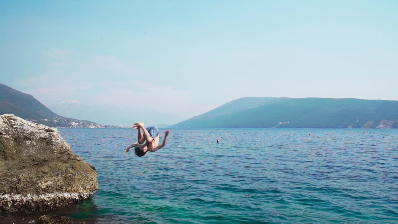 Caucasian young boy jumping into sea from a rock in slow motion doing somersault