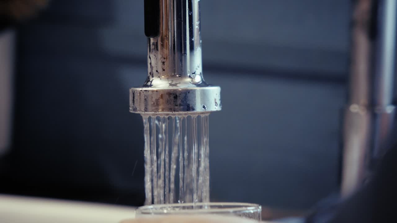 Person filling a clear glass with tap water, symbolizing hydration and daily routine, dominant clear and metallic tones in a kitchen setting