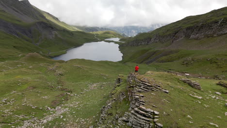 Circular-aerial-shot-of-a-young-man-with-a-red-jacket-Bachalpsee-lake-in-Switzerland-on-a-cloudy-day-with-some-traces-of-sunlight