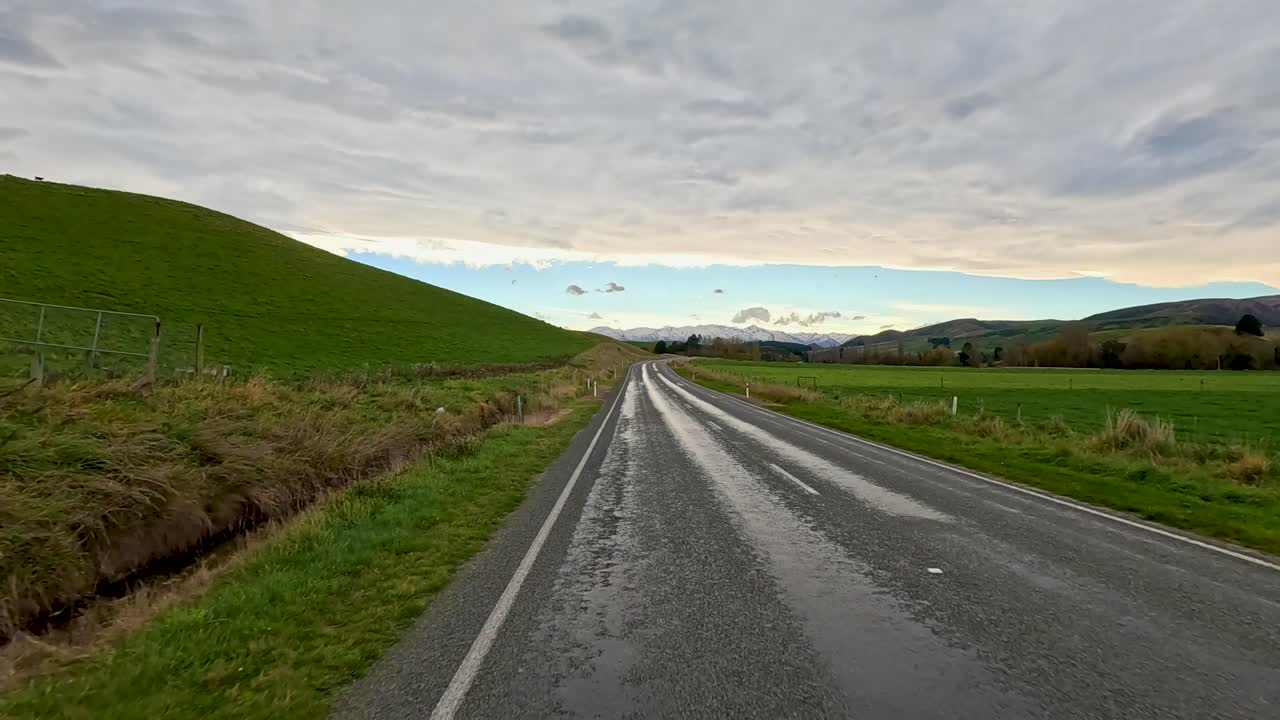 Forward-moving timelapse along a wet rural road bordered by grassy hills under overcast skies, captured with smooth camera motion and natural daylight