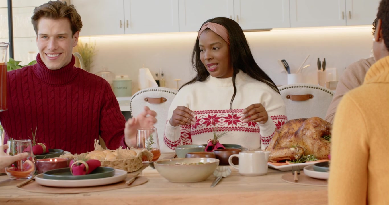 Diverse group at table after toast, man in red sweater pouring pitcher for Christmas drinks, turkey