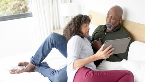 Feliz-Pareja-Madura-Y-Diversa-Sentada-En-La-Cama-Usando-Una-Tableta-Y-Hablando-En-Casa,-Cámara-Lenta