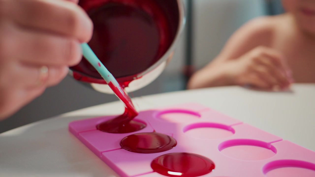 Mano vierte líquido rojo vibrante en un molde de silicona rosa. Un niño observa con curiosidad desde atrás. El líquido brillante llena cada cavidad lentamente. Ideal para manualidades culinarias, proyectos de bricolaje