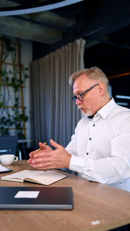 busy adult caucasian male in white shirt sitting at desk writing in the notebook. businessman talks to somebody with irritation. vertical video.