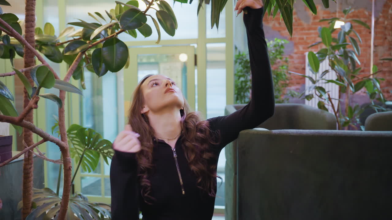 Young woman reaches toward large green leaves of indoor plant with natural curiosity while standing near window as blurred vehicle motion adds depth to calm interior filled with greenery lighting