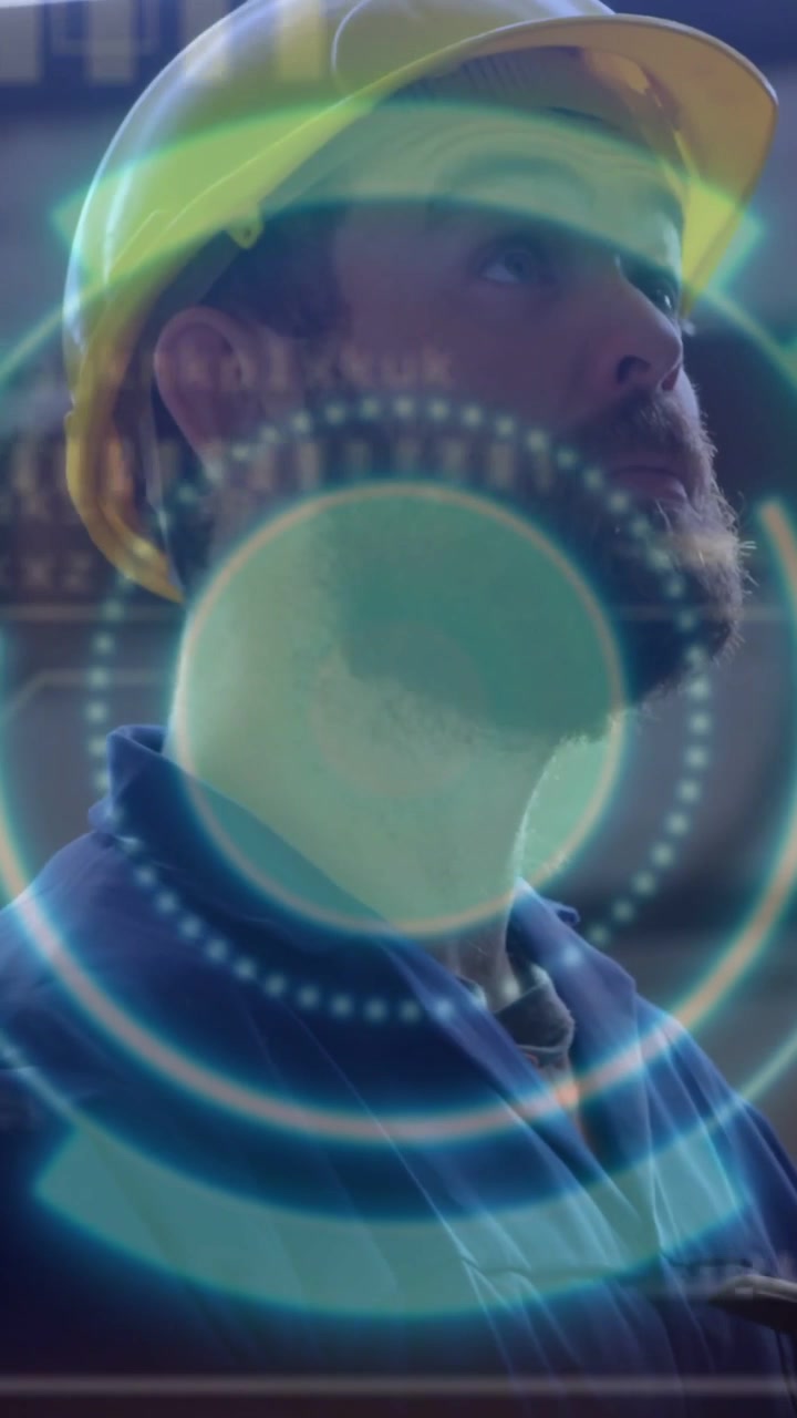 Video verticale: Uomo con elmetto giallo e camicia blu che usa un tablet come HUD pulsante, guardando in alto per un'ispezione.