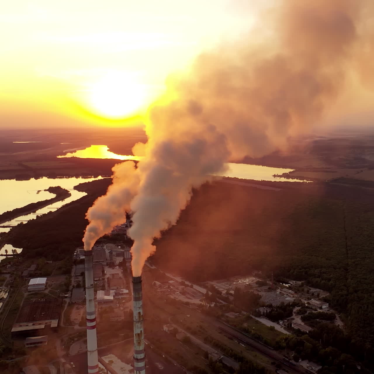 Pipes with smoke on the natural green fields background. Clouds of smoke coming from the chimneys of the plant with yellow lighting of setting sun. Aerial view.