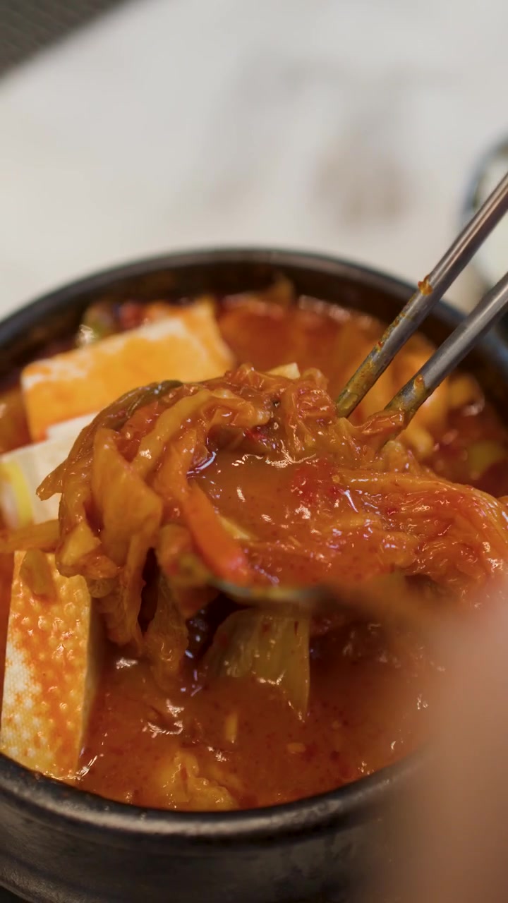 Pengambilan gambar jarak dekat seseorang menggunakan sumpit logam untuk makan sup kimchi Korea pedas dengan tahu