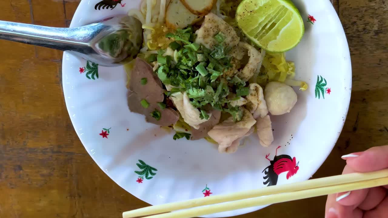 A close-up view of noodles being mixed with lime, herbs, and chopsticks in a decorative bowl.