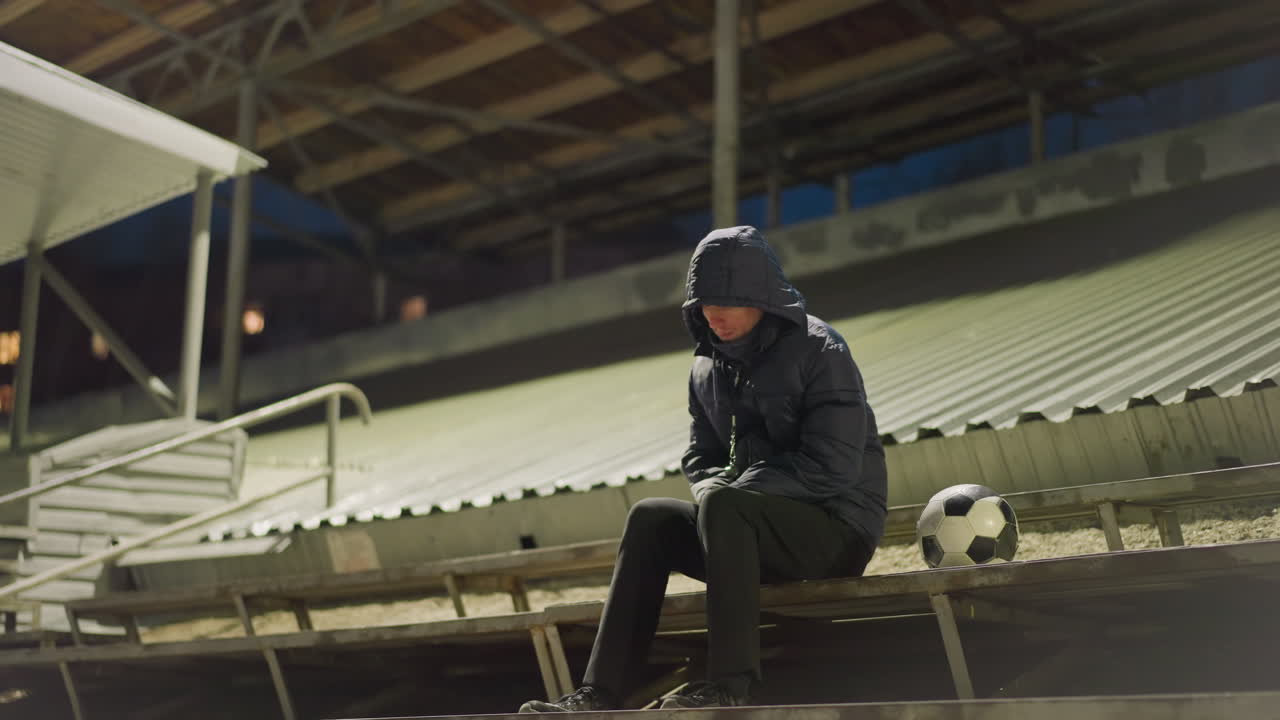 un hombre vestido con un traje negro y una chaqueta se sienta en silencio y solo en un estadio débilmente iluminado, con las manos entre las piernas, con una pelota de fútbol cerca