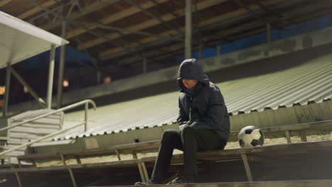 a man dressed in a black outfit and jacket sits quietly and alone in a dimly lit stadium, his hands clasped between his legs, with a soccer ball nearby