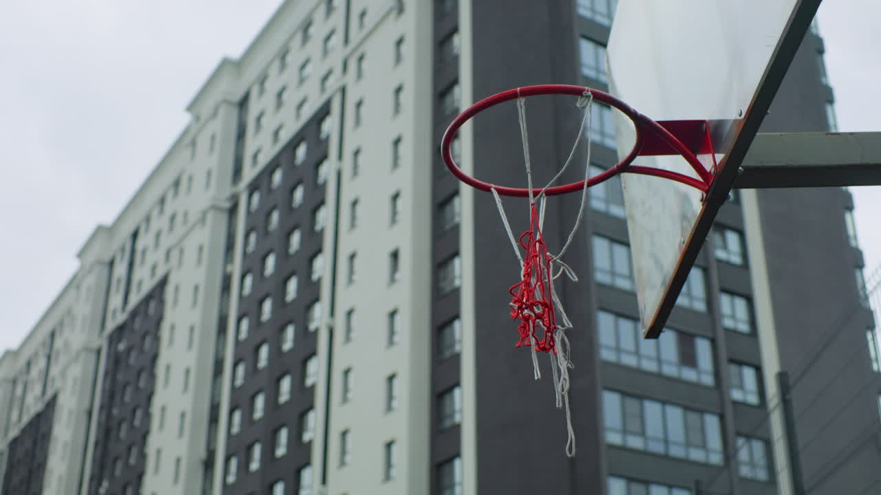 canestro da basket urbano su sfondo di grattacieli con bordo in metallo rosso e rete a catena sfilacciata, cielo nuvoloso, imponenti condomini, campo in cemento grezzo, colori tenui, atmosfera di solitudine urbana, nostalgico