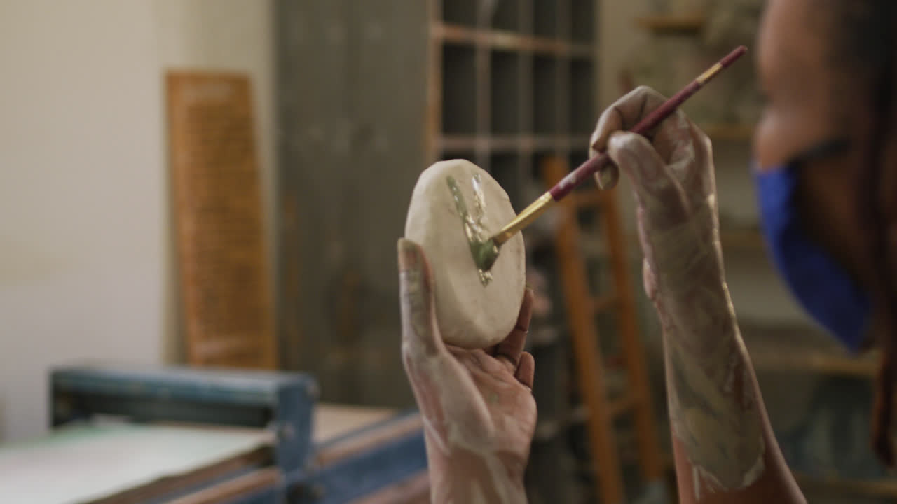 여성 도자기가 얼굴 마스크와 앞옷을 착용하고 도자기 스튜디오에서 도자기에 을 그리기 위해 브러쉬를 사용합니다.