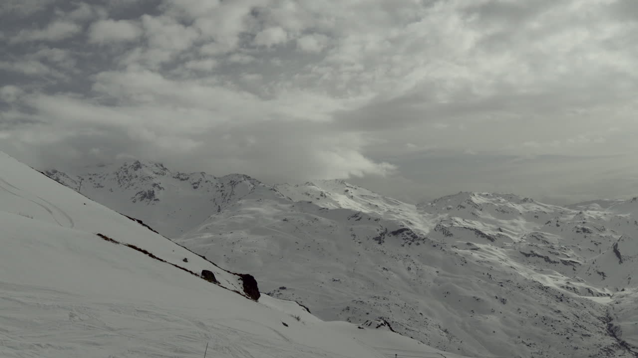 Timelapse of snowy alpine mountain peaks under a cloudy sky. Clouds drift dynamically across the rugged winter landscape. Slopes and ridges covered in fresh snow. Three Valleys, France