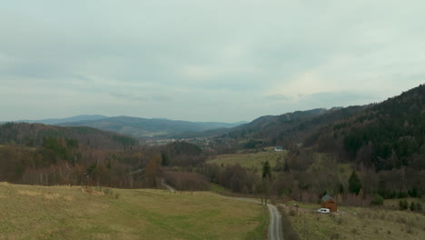 Aerial---winding-dirt-road-leads-to-a-solitary-house-nestled-in-a-lush-mountainous-region,-with-a-small-village-in-the-distance-under-an-overcast-sky