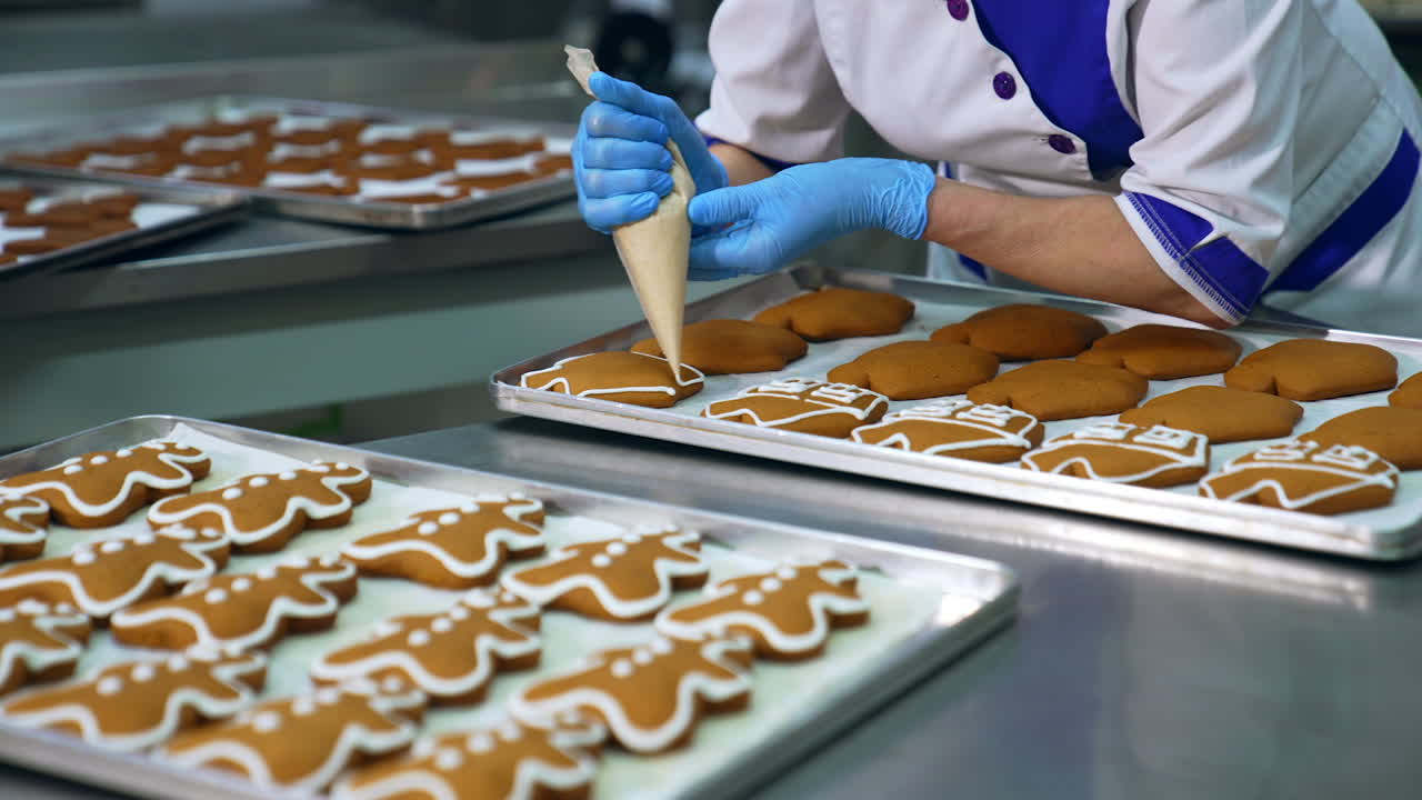 Worker squeezing icing cover on cookies from a pastry bag. Decorating gingerbread sweets on a confectionery factory. Trays with cookies in blur at background.