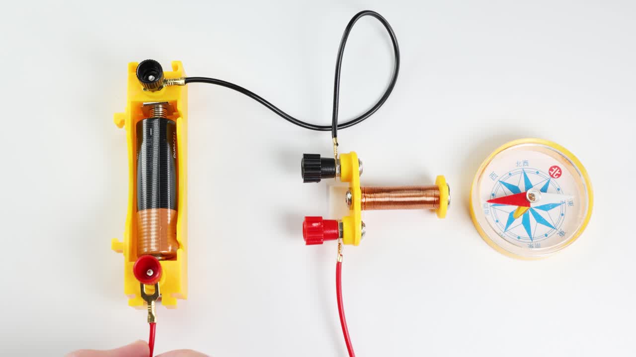 Hands demonstrate magnetic force using a battery, coil, and compass on a white background