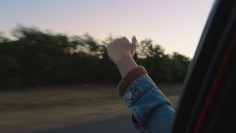 Frau-Im-Auto-Hält-Die-Hand-Aus-Dem-Fenster-Und-Spürt,-Wie-Der-Wind-Durch-Die-Finger-Weht,-Während-Sie-Im-Sommerurlaub-Auf-Dem-Land-Unterwegs-Ist-Und-Bei-Sonnenuntergang-Die-Freiheit-Auf-Der-Straße-Genießt