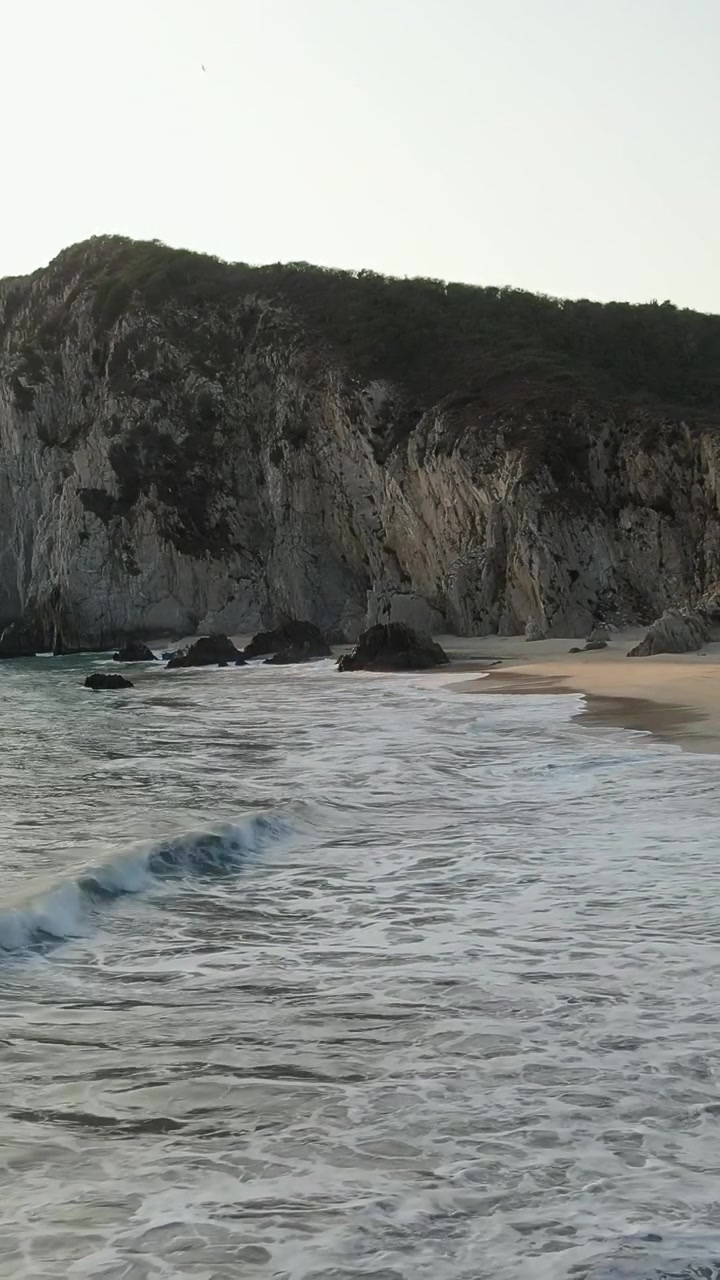 Ripresa aerea verticale statica delle onde del Pacifico che si infrangono sulla spiaggia di Maruata con sabbia bianca e scogliere aspre al tramonto.