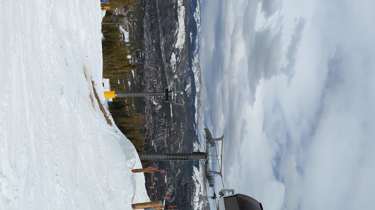 Seggiovia che trasporta sciatori e snowboarder in cima a una pista da sci in una giornata nuvolosa, verticale.