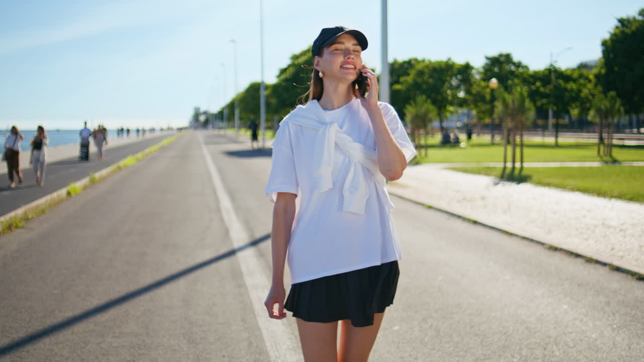 Smiling lady speaking cellphone strolling asphalt road in black cap. Happy girl