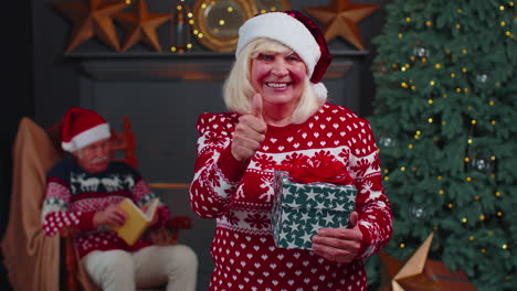 Positive-senior-grandmother-in-Christmas-sweater-smiling-friendly-at-camera-and-raises-thumbs-up