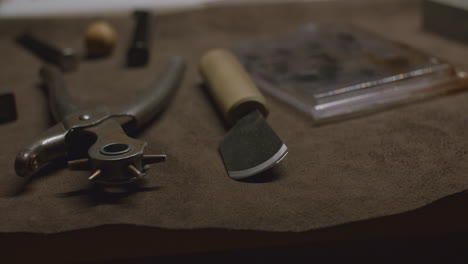 a skiving knife with a wooden handle and a rotary hole punch tool resting on a brown suede work mat, used in leathercraft.