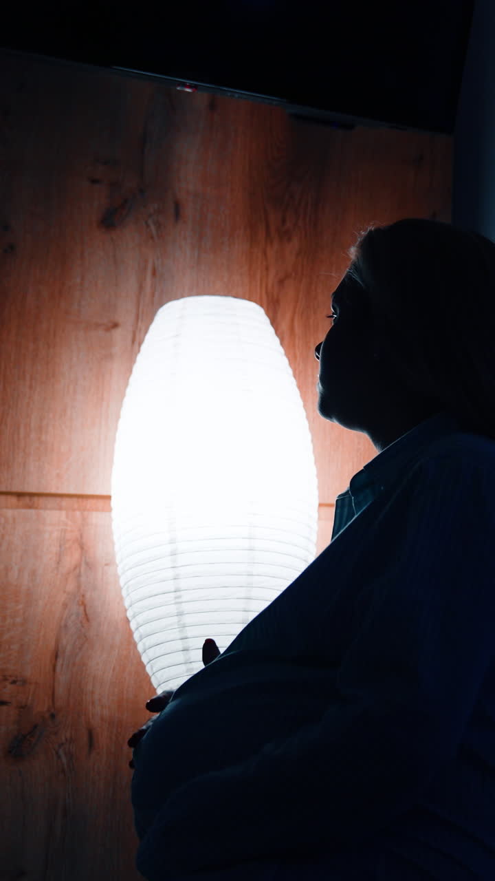 Silhouette of pregnant female sitting on the bed holding by the belly. Worried future mom in a dark room. Vertical video